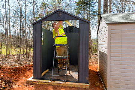 Worker in reflective vest puts finishing touches at storage shelter while standing on ladderの写真素材