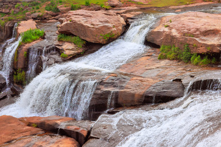 Tranquil waterfall flows over stones surrounded by lush greenery under bright sunlight.の写真素材