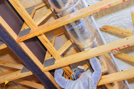 Construction worker is fitting thermal barrier insulation material above wooden ceiling frame in attic house being built.の写真素材