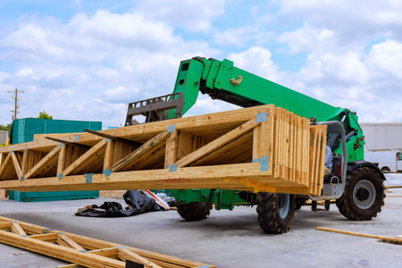 Construction vehicle is lifting large wooden truss beams at job site under during works daytime.