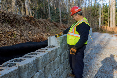 Construction worker measures stone blocks while working on retaining wall beside gravel road