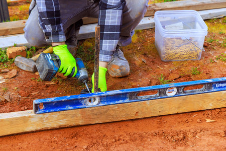 Contractor in gloves drills into wood while using level to ensure accuracy in backyard construction...
