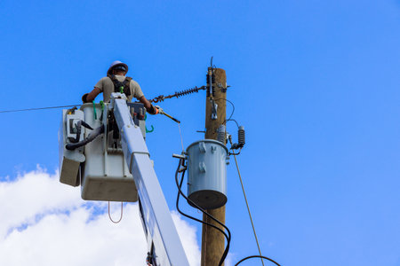 Utility worker inspects electrical equipment atop bucket truck under bright sunlight few clouds.