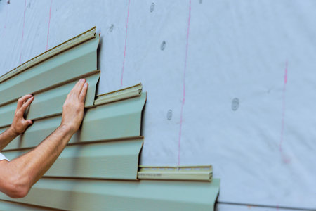 Workers are carefully aligning securing green PVC vinyl siding panels on exterior of home under...