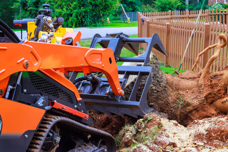Workers are using compact track loader to extract large tree stump from backyard in suburban...