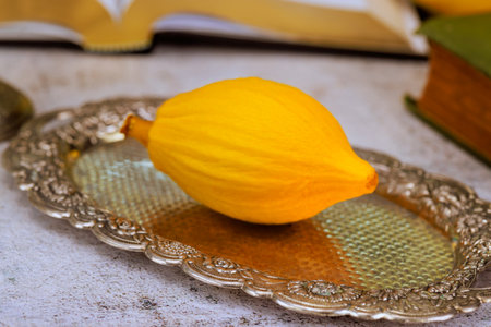 During Hebrew tradition yellow etrog rests on an ornate silver platter, surrounded by aged books,...