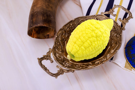 Yellow etrog in an ornate metal dish beside shofar, tallit gold stripes, decorative kippah on light marble surface.の写真素材