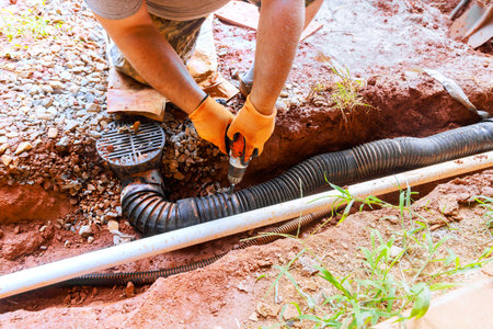 Contractor works on fixing drainage pipe in yard, wearing gloves using power drill tool on work day.の写真素材