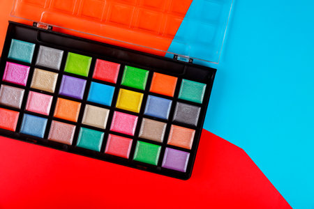 Bright eyeshadow palette with numerous colors is placed on bold background of red, blue shapes.
