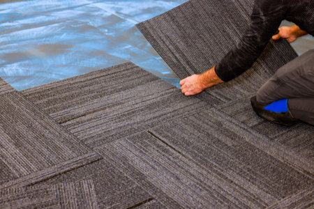 Worker is carefully laying down grey carpet tiles on blue protective covering