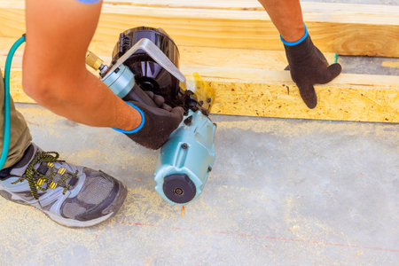 Carpenter in gloves operates pneumatic nail gun to secure wooden boards on floor of busy construction site.の写真素材