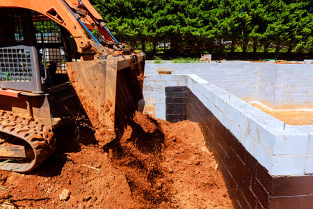 Construction worker operates excavator to load soil into building foundation