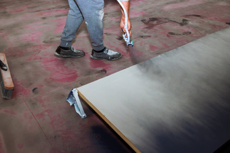 Worker is applying paint to wooden board using spray gun while standing on dusty floor