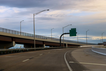 Cars travel on curved highway below large overpass with street lamps on cloudy day.