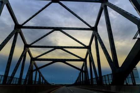Person drives under large metal bridge with clouds above in evening light.の写真素材