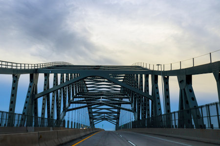 Metal bridge spans across road with gray clouds above clear path for vehicles below at dusk.