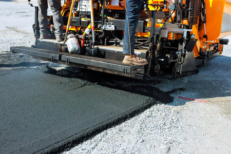 Construction workers operate an asphalt paver to apply fresh pavement on city road