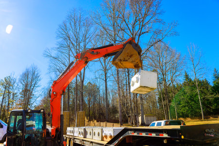 Large excavator lifts cement block from flatbed truck at construction site