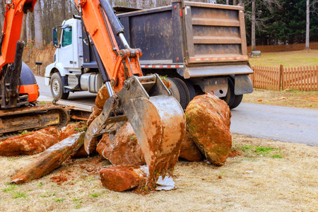 Digger uses its claw to lift large rocks from ground in residential area while truck waits close by.