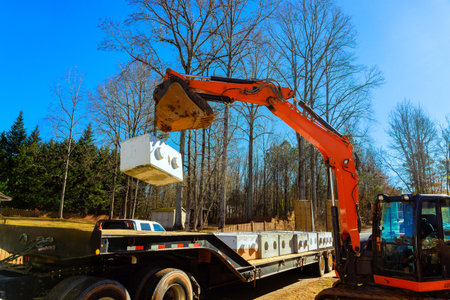 Worker uses excavator machine to move large concrete blocks from truck in construction area