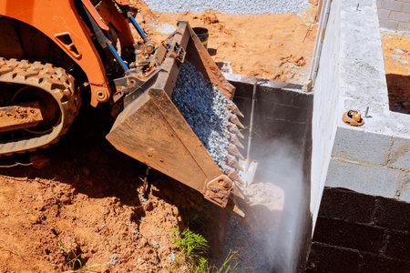 Construction excavator bucket dumps gravel near foundation of building while work continues site.