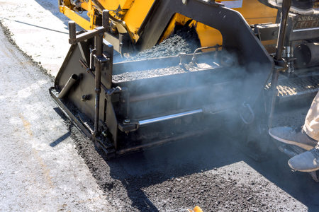 Paving machine is laying fresh asphalt on road workers are machine emits smoke into air.
