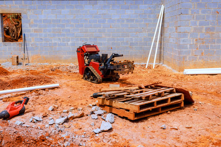 Small machine works in dirt at construction site with pallets pipes nearby