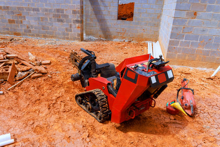 Small construction digging machine sits on dirt surface at building site with bricks pipes nearby