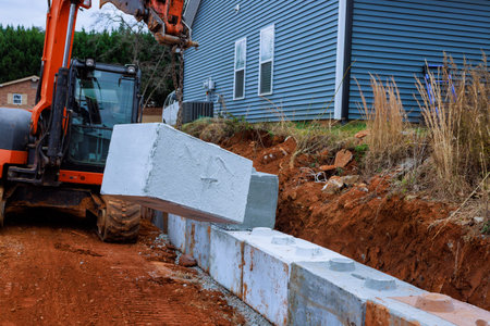 Construction workers use excavator to lift place concrete blocks for retaining walls beside house.