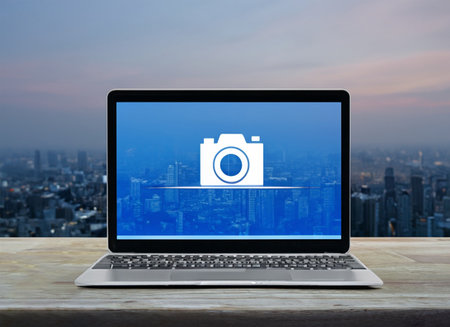 Camera flat icon on modern laptop computer screen on wooden table over modern city tower and skyscraper, Business photography online conceptの素材