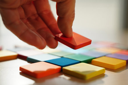 Hand arranges tiles. Puzzle game concept. Colorful pieces on tabletop. Creative studio setting.の素材