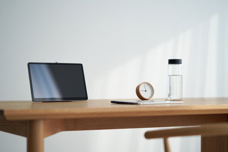 Tablet, clock, bottle, stationery on table. Brightly lit room, workplace, office.の素材