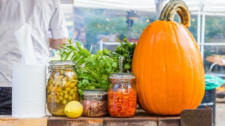 Glass jars with fresh food, herbs and a large pumpkin with a man cooking in the backgroundの写真素材