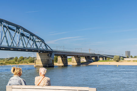 Two woman sitting on bench enjoying the view from the river quay called Waalkade in Nijmegen The Netherlands on a sunny autumn day.の写真素材
