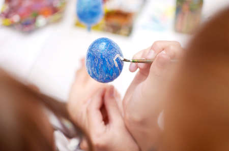 Child hands painting white lines on blue Easter egg with brush