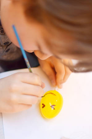 Child hands painting smiley face on yellow Easter egg with brushの写真素材