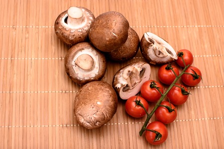 Gray mushrooms champignons and red cherry tomatoes lie on a beech wooden mat.の写真素材