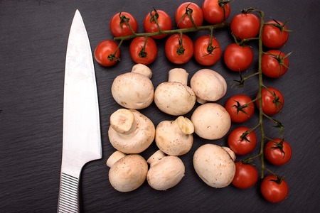 Red cherry tomatoes and champignon mushrooms, kitchen knife lie on a black stone blackboard, background.の写真素材