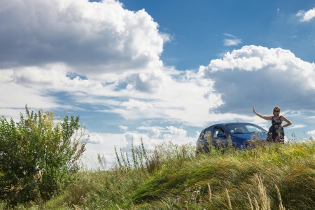 Woman standing near a new car in a field under blue sky with white cloudsの写真素材