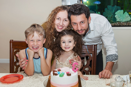 Happy family of four celebrating birthday of happy little girl sitting at the table with birthday cake on itの写真素材