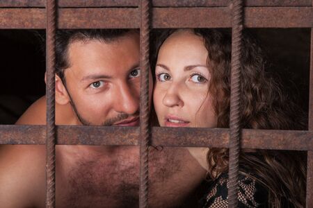 Portrait of a young couple in jail, loving man and woman looking out of rusty iron rodsの写真素材