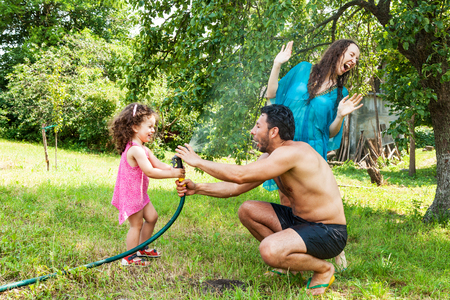 Dad and mom play with a little daughter outdoors in the summer, pouring water from a hoseの写真素材