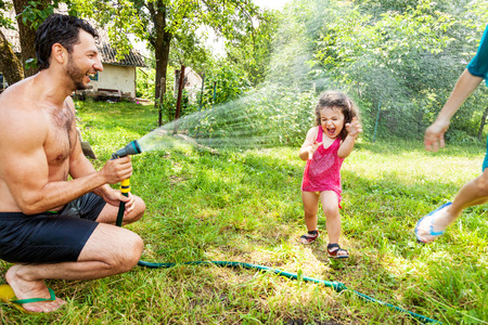 Dad and mom play with a little daughter outdoors in the summer, pouring water from a hoseの写真素材