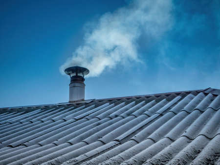 Stainless steel chimney on a slate roof emits smoke against a blue skyの写真素材