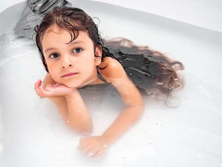 Baby girl with long hair in a mermaid costume lying in the water in the bathtub and looks into the...