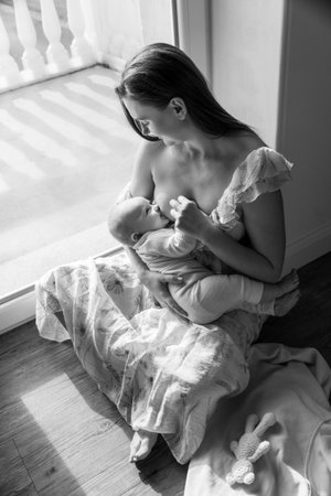 A woman breastfeeds her baby sitting on the floor by the window. Kid sucks breast milk. caring tenderness protection. mother love concept. natural nutrition., monochrome photoの写真素材