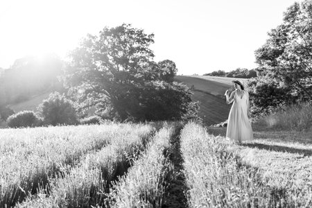 A pregnant woman in a straw hat stands on the edge of a blooming lavender field in the rays of the setting sun, monochrome imageの写真素材