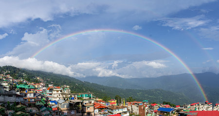 Vibrant Kalimpong town with colorful rooftops stretches along the valley as a dramatic double rainbow arches across a bright sky.の写真素材