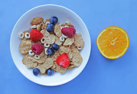 Bowl of breakfast cereals with berries and an orange on blue table.