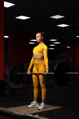Young beautiful sportive girl in a yellow tight-fitting body tracksuit does deadlift in the gym....